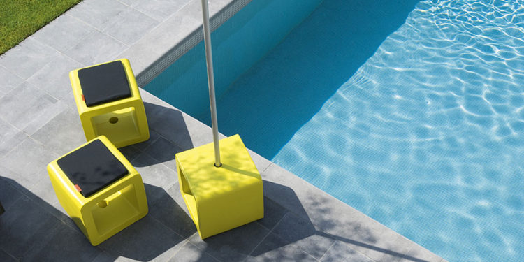 trois exemplaires d'un cube au bord d'une piscine, l'un servant de base à un parasol, deux autres de pouf avec un coussin dessus